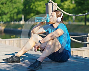 Exhausted young sportive man in headphones drinking water from bottle during workout outdoors