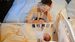 Exhausted young woman rocking crib of her sleepless newborn baby. Maternal depression after childbirth and sleepless