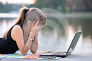 Exhausted young woman working behind laptop computer lying down in summer park outdoors. Problems with remote work concept