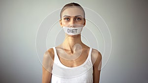 Exhausting diet concept, miserable slim woman with taped mouth looking at camera