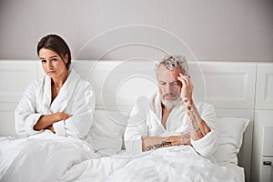 Exited brunette woman and thoughtful bearded man in white bathrobes looking away in bedroom