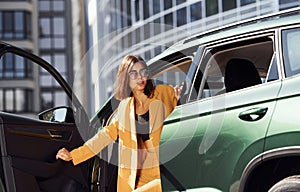 Exits vehicle. Young fashionable woman in burgundy colored coat at daytime with her car