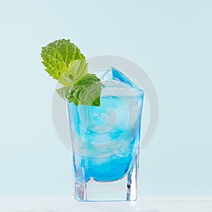 Exotic alcohol cold blue cocktail with curacao liquor, ice and green mint in rich wet shot glass on soft light pastel green.