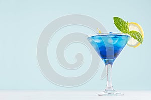 Exotic cold shot glass cocktail with blue curacao, ice cubes, lemon slice, yellow straw, green mint on soft light mint color.