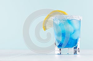 Exotic cold shot glass cocktail with blue curacao, ice cubes, salt rim, lemon slice on soft light mint color background.