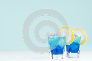 Exotic cold shot glass cocktail with yellow lemon slice, blue curacao, ice cubes, salt rim on soft light mint color background.