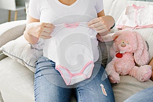 The expectant mother holds a baby white bodysuit with a pink canticle in her hands, and a pink stuffed toy hare is sitting next to