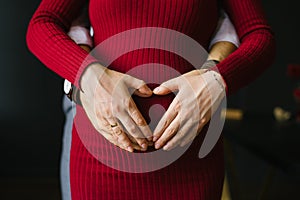 Expecting a baby. Hands of husband and wife on pregnant belly in red dress.