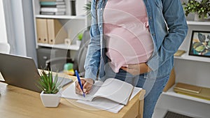 Expecting joy, young, hispanic working-mum-to-be massages belly while taking notes at office