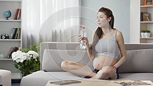 Expecting woman sitting on sofa drinking water keeping water balance, healthcare