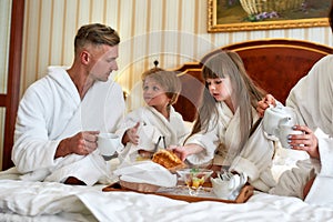 Experience more together. Parents and two kids in white bathrobes having breakfast in bed, eating pastries and drinking