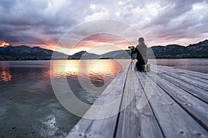 Experience nature with the hobby of outdoor photography - A man kneeling on a wooden pier of a lake adjusts his camera aiming at a