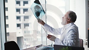 Experienced adult doctor with white beard siting at desk and examining X-ray