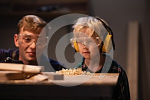 An experienced carpenter and a small boy in safety glasses blow sawdust off a work table in a carpenter`s workshop.