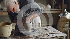 Experienced ceramist grey-haired bearded man is smoothing molded ceramic pot with wet sponge. Spinning throwing wheel