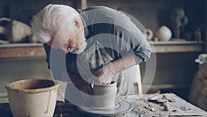 Experienced ceramist grey-haired bearded man is smoothing molded ceramic pot with wet sponge. Spinning throwing wheel