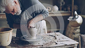 Experienced ceramist grey-haired bearded man is smoothing molded ceramic pot with wet sponge. Spinning throwing wheel