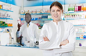 Experienced female pharmacist standing on background with male colleague in pharmacy