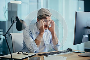 An experienced gray-haired businessman works at the computer until late, tired has a severe headache, stress and depression