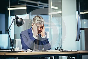 Experienced gray-haired businessman works at the computer until late, tired has a severe headache, stress and depression