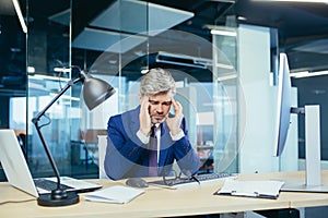 Experienced gray-haired businessman works at the computer until late, tired has a severe headache, stress and depression