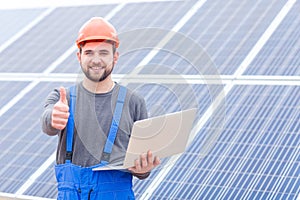 Experienced worker of the solar battery station holds a laptopand shows a thumbs-up gesture.