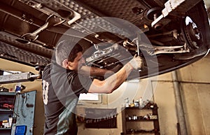 Experienced young car mechanic standing under lifted car during repair process in garage. Car check up and maintenance