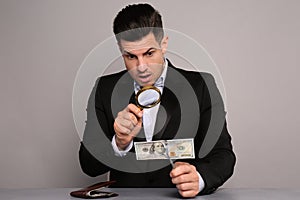 Expert authenticating 100 dollar banknote with glass at table on light grey background. Fake money concept