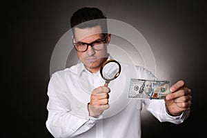 Expert authenticating 100 dollar banknote with magnifying glass against dark background, focus on hand. Fake money concept