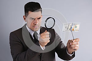 Expert authenticating 100 dollar banknote with magnifying glass on light grey background. Fake money