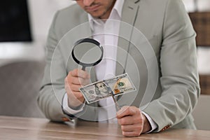 Expert authenticating 100 dollar banknote with magnifying glass at table in office, closeup. Fake money concept