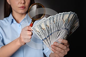 Expert authenticating 100 dollar banknotes with magnifying glass against dark background, closeup. Fake money concept