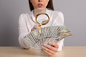 Expert authenticating 100 dollar banknotes with magnifying glass at table on light grey background, closeup. Fake money concept