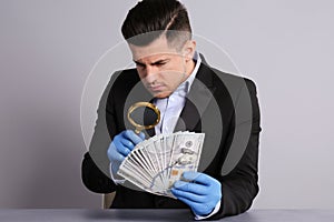 Expert authenticating 100 dollar banknotes with magnifying glass at table on light grey background. Fake money concept