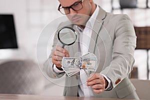 Expert authenticating 100 dollar banknotes with magnifying glass at table in office, focus on hand. Fake money