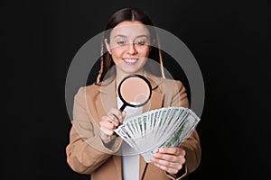 Expert authenticating 100 dollar with magnifying glass against black background, focus on hand. Fake money concept