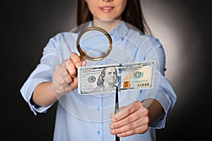 Expert authenticating 100 dollar with magnifying glass against dark background, closeup. Fake money concept