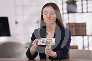 Expert authenticating 100 dollar with magnifying glass at table in office, focus on hand. Fake money concept