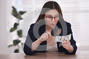 Expert authenticating dollar banknote with magnifying glass at table in office, focus on hand. Fake money concept