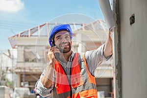 An expert young engineer speaks on the phone. Wears a blue helmet and an orange construction vest. Engineer working on