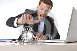 Exploited businessman at office desk stressed and frustrated with alarm clock in out of time and deadline concept