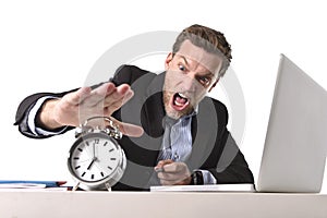 Exploited businessman at office desk stressed and frustrated with alarm clock in out of time and deadline concept