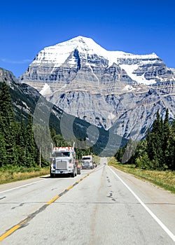 Two tree transportating trucks transport on the canadian highway in front of a snowy mountain