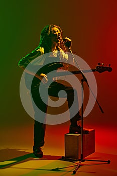 Expressive musician with dreadlocks playing electric guitar and singing on microphone on stage against gradient red