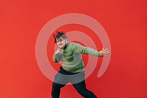 Expressive overweight young man in stylish casual clothes dancing hip hop on red wall background. The fat man shows a dance
