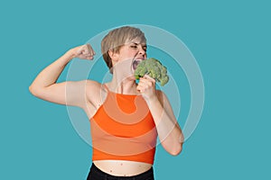 An expressive portrait of a young woman with broccoli as dumbbells on a blue background. Healthy diet, healthy lifestyle