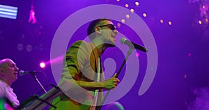 Expressive singer men in glasses singing into microphone. Live music concert in front of bright colorful strobing lights