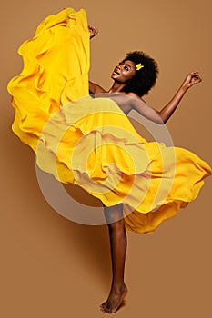 Expressive Woman dancing in Yellow Flying Dress. Happy Dark Skinned Dancer in Waving Fabric Gown. Model with Black curly Afro Hair