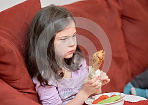 expressive young girl is eating chicken and vegetables for dinner on the couch while watching cartoons