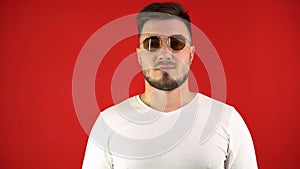 Expressive Young Man on a Red Background Puts on Fashionable Sunglasses, Macho Man. Studio Portrait of a Handsome Man